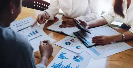 Grupo de profissionais analisando relatórios financeiros, gráficos e indicadores contábeis sobre uma mesa durante reunião de gestão empresarial.