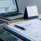 Mesa de trabalho com documentos fiscais organizados, laptop com dados financeiros ao fundo, calculadora e calendário, representando revisão contábil e planejamento tributário.